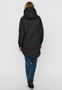 Cappotto lungo imbottito nero con cappuccio, caratterizzato da un motivo a rombi, polsini elasticizzati e orlo dritto, abbinato a jeans blu e scarpe nere.