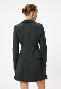 Blazer gris a medida con cuello y falda plisada. El material parece texturizado, con una silueta ajustada y líneas limpias.