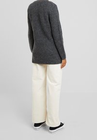 Grå stickad tröja med långa ärmar, bärs över krämfärgade kordflöjlsbyxor med vida ben. Svarta sneakers med vita sulor kompletterar outfiten.