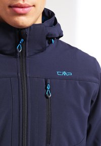 Veste softshell marine avec col montant, fermetures éclair bleues et logo CMP. Comprend des poches poitrine et latérales, des coutures élégantes et une texture lisse.