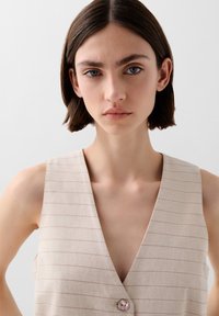 Beige vest met verticale strepen, V-hals en een enkele knoopsluiting met een ronde, iriserende detail. Vloeiende textuur.