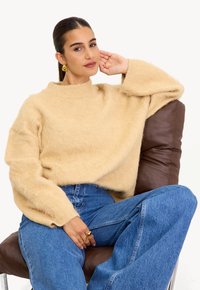 Femme aux cheveux noirs portant un pull moelleux beige et un jean bleu, assise sur une chaise en cuir marron, la tête posée sur la main, portant des boucles d'oreilles et des bagues en or.