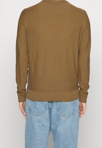 Pull en maille marron avec un col rond et des poignets côtelés. Porté avec un jean bleu clair, montrant les détails du dos et du côté du vêtement.