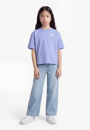 Ung person står iført en lilla Nike-t-shirt, lyseblå vidde-bukser og hvide sneakers mod en ensartet hvid baggrund.