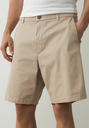 Beige knee-length shorts with front button and zipper, belt loops, side pockets; worn with white tucked-in shirt.