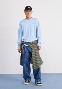 Camisa de manga longa azul clara com o texto "nike", camisa às riscas verde-azeitona amarrada na cintura, jeans em estilo solto, sapatilhas pretas e brancas.