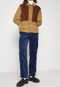 Manžestrová bunda s hnědým horním panelem a béžovým spodním panelem, doplněná modrými manžestrovými kalhotami a černými botami. Má patentní knoflíky a límec.