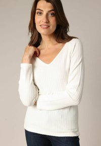 Pull blanc à manches longues avec un col en V, présentant une texture côtelée et de fins motifs verticaux. Porté sur un jean foncé.