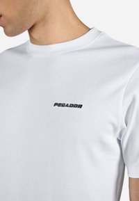 Biely bavlnený tričko s okrúhlym výstrihom a jemne čierne vyšívané logo "PEGADOR" na ľavej hrudi. Hladká textúra.