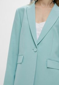 Blazer azzurro chiaro con tessuto texturizzato, revers a punta, chiusura con un bottone e due tasche frontali con pattina.