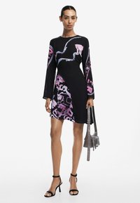 Schwarzes Kleid mit langen Ärmeln, das pinke und blaue abstrakte Muster aufweist. Es hat eine figurbetonte Silhouette und wird mit schwarzen Riemchen-High Heels und einer grauen Tasche kombiniert.