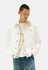 Giacca bianca con colletto in corduroy beige, bottoni a pressione dorati, abbinata a un gilet testurizzato beige e una camicia bianca, indossata con jeans blu.