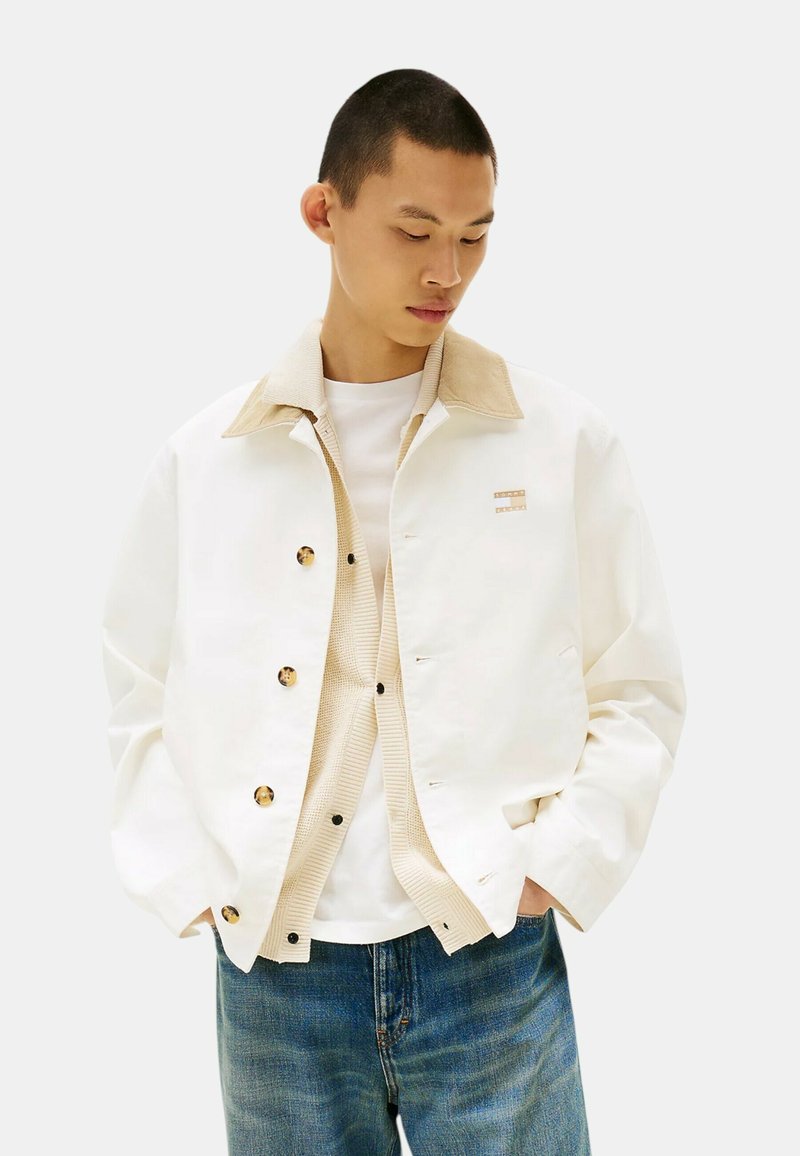 Giacca bianca con colletto in corduroy beige, bottoni a pressione dorati, abbinata a un gilet testurizzato beige e una camicia bianca, indossata con jeans blu.