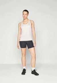 Νεαρή γυναίκα που στέκεται φορώντας ανοιχτό ροζ αμάνικο, μαύρες σορτς ποδηλασίας, λευκές κάλτσες και μαύρα αθλητικά παπούτσια μπροστά από ένα απλό φόντο.