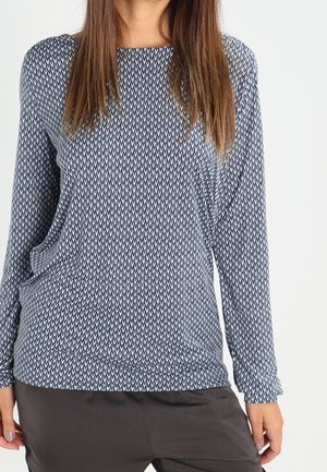 Vrouw draagt een top met lange mouwen in marineblauw en wit met een patroon en een ronde hals, gecombineerd met donkergrijze broek.