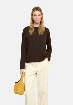 Kobieta ubrana w ciemnobrązową bluzkę z długim rękawem i kremowe spodnie ze sztruksu, trzymająca małą zamszową torebkę w kolorze musztardowym z krótkim uchwytem.