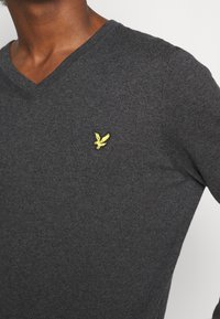 Suéter gris de cuello en V hecho de tejido de punto, con un pequeño logotipo de un pájaro amarillo bordado en el pecho. Ajuste clásico con superficie texturizada.