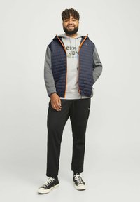 Mørkeblå quilted vest med orange lynlås, grå hættetrøje, sorte joggingbukser og sorte sneakers. Løst lag-på-lag look med hætte.