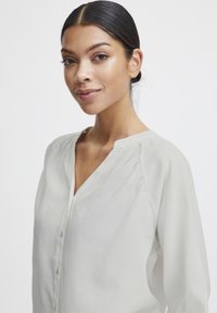 Blusa grigia chiaro con scollatura a V, pieghe sottili e chiusure a bottoni. Realizzata in un tessuto liscio con una texture morbida.