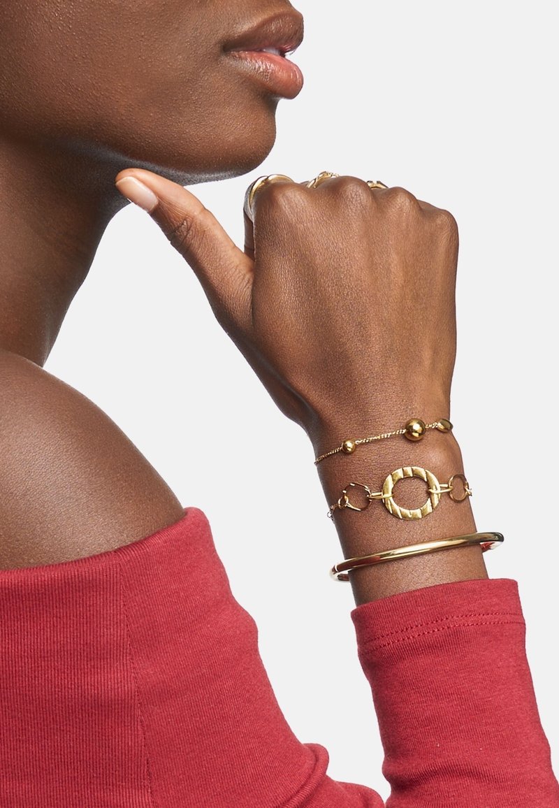 Gold bracelets adorn a hand with textured accents, featuring a circular link, smooth and beaded designs, against a red fabric background.