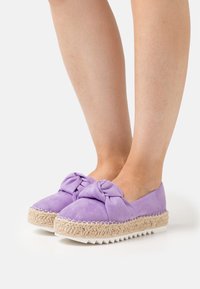 Nohy v fialových semišových espadrilích na platformě s uzlem a tkanou jutu střední podrážkou na bílém pozadí.