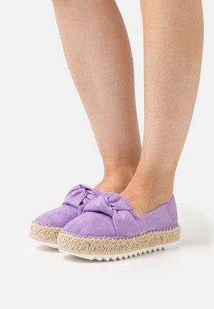 Nohy v fialových semišových espadrilích na platformě s uzlem a tkanou jutu střední podrážkou na bílém pozadí.