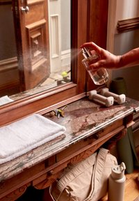 Eine klare Glas-Sprühflasche in der Hand, die eine Flüssigkeit in Richtung eines Spiegels sprüht. Marmorarbeitsplatte mit einem weißen Handtuch, Schlüsseln und Hanteln in der Nähe.