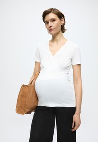 Λευκή τοπ για εγκύους τύπου wrap, με κοντά μανίκια και εφαρμοστό σχέδιο. Συνδυασμένο με καφέ φελτ τσάντα. Μαύρο παντελόνι ορατό από κάτω.