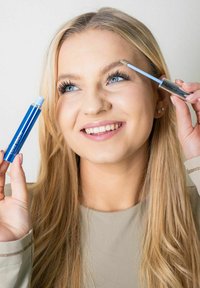 Blå maskara rør i den ene hånden og en klar bryngele-applikator i den andre. Har langt hår og en nøytral-farget topp. Minimal bakgrunn.