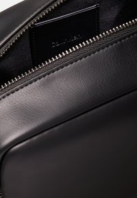 Hoek van een zwarte leren tas met een gestructureerd oppervlak, metalen ritssluiting en binnenzak. Ritstrekker en merkteken zichtbaar aan de binnenkant.