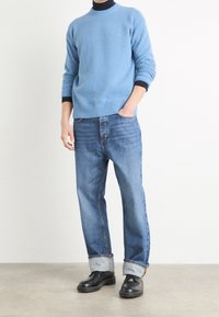 Ljusblå stickad tröja med ribbade muddar och nederkant, kombinerad med ljusblå jeans och svarta läderskor, står på en neutral bakgrund.