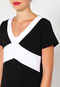 Czarna sukienka z krótkimi rękawami i głębokim dekoltem w kształcie litery V, z białym przekątnym pasem na wysokości klatki piersiowej. Gładki materiał o prostym fasonie.