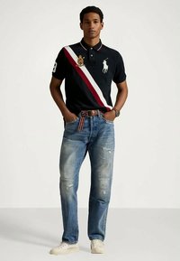 Marineblå poloshirt med hvite og røde diagonale striper, logodetalj, kombinert med lyseblå ripped jeans og beige sneakers.