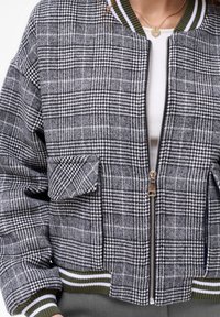 Szara i biała bomberka w kratę z zamek błyskawiczny z przodu, kieszeniami na łatki i ściągaczami w kolorze zielonym i białym. Teksturowana tkanina.