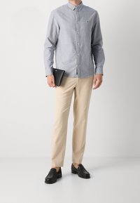 Muž v svetle modré košili na gombíky a béžových kalhotách, který drží černou peněženku, stojí proti jednoduchému pozadí.