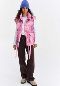 Różowa pikowana kamizelka w kwiaty z wysokim kołnierzem, różowymi troczkami i przednimi kieszeniami. Nosi się ją na białym topie, w połączeniu z brązowymi spodniami o szerokich nogawkach i beżowymi butami.
