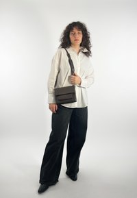 Borsa a tracolla in pelle marrone con chiusura a patta e dettagli testurizzati, indossata da una persona con camicia bianca e pantaloni neri a gamba larga.