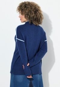 Frau mit lockigem Haar, die einen blauen Pullover mit hellblauen Details trägt, steht mit hinter dem Rücken verschränkten Händen nach links gerichtet vor einer weißen Wand.