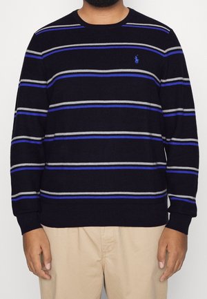 Homme portant un pull noir avec des rayures horizontales bleues et grises et un pantalon beige, debout devant un fond blanc uni.