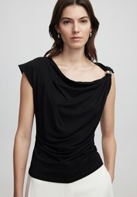 Černý top s jedním ramínkem a stříbrným akcentem na rameni. Měkká textura a volný střih, doplněný světlými kalhotami.