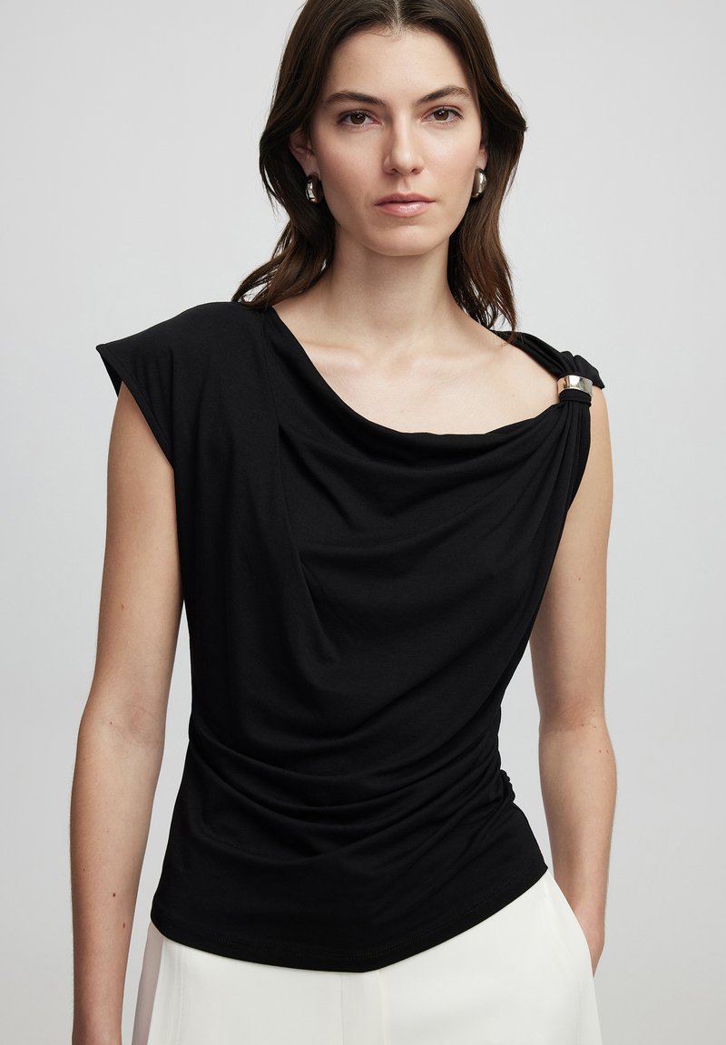 Černý top s jedním ramínkem a stříbrným akcentem na rameni. Měkká textura a volný střih, doplněný světlými kalhotami.