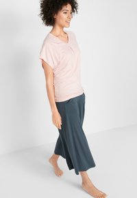 Top rosa chiaro con scollo a V e maniche corte arrotolate, abbinato a pantaloni grigi scuri, fluidi, lunghi fino al polpaccio. Tessuto morbido con vestibilità rilassata.