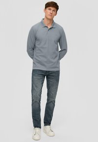 Langärmliges graues Poloshirt mit einer Knopfleiste aus zwei Knöpfen, kombiniert mit dunkelblauen Jeans und weißen Sneakers, vor einem neutralen Hintergrund stehend.