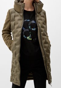 Oliwkowa ocieplana kurtka z wysokim kołnierzem, zamkiem błyskawicznym i kieszeniami bocznymi. Nosi się ją na czarnej bluzie z grafiką w okrągłym kształcie.
