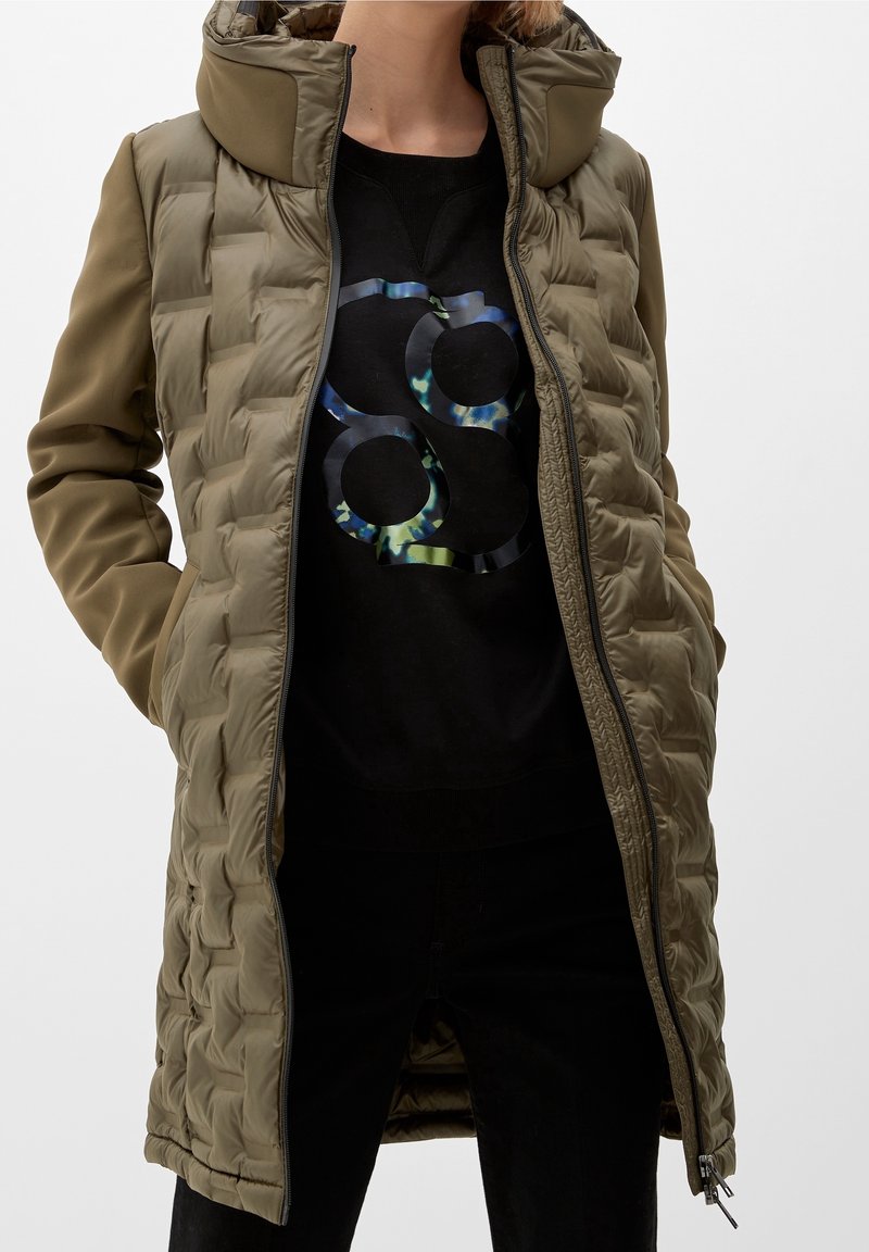 Oliwkowa ocieplana kurtka z wysokim kołnierzem, zamkiem błyskawicznym i kieszeniami bocznymi. Nosi się ją na czarnej bluzie z grafiką w okrągłym kształcie.