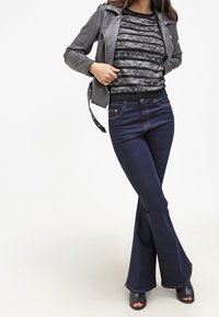 Veste en cuir noire, haut à motif en dentelle, jean évasé bleu foncé, et mules à talons noires, avec une texture lisse et une coupe ajustée.