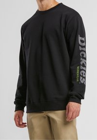 Mustapaita, jossa on pitkät hihat, vasemmassa hihassa harmaa "Dickies" -logo ja vihreä "WORKWEAR" -teksti. Pehmeä rakenne, klassinen malli.