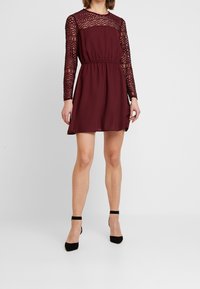 Robe bordeaux avec manches longues en dentelle, encolure ronde, taille cintrée et ourlet fluide, assortie à des chaussures à talons noires avec bride à la cheville.