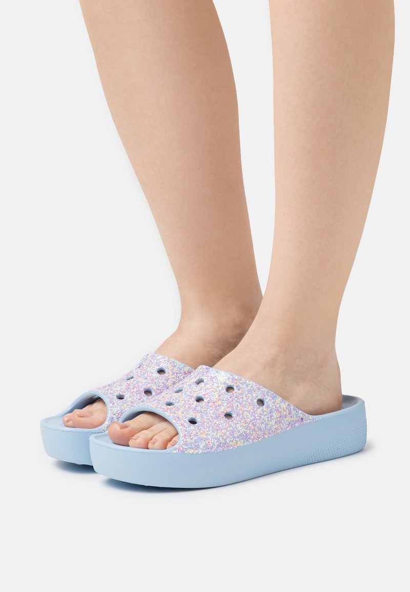 Sabots violets scintillants à enfiler avec des trous découpés, une base bleue et une semelle texturée pour l'adhérence. Les pieds sont visibles, montrant les orteils et la voûte plantaire.