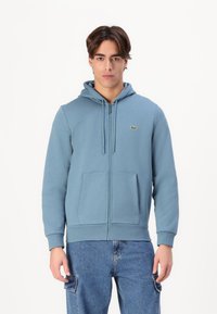 Haut de sweat à capuche zippé bleu avec une poche avant, capuche à cordon de serrage et logo sur la poitrine. Fabriqué en tissu doux et texturé. Design décontracté.